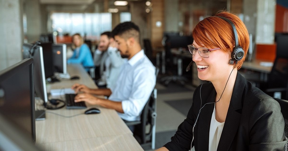 red haired call center agent smiling at desk with other agents lined up behind