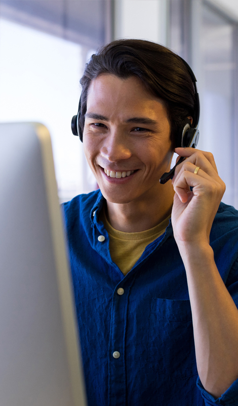 man in a blue shirt sitting at a computer talking to someone on a headset